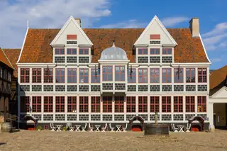 Half-timbered Mintmaster House with red-framed windows, twin gables and chairs on the cobbled square.