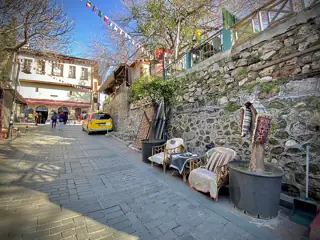A paved lane passes a stone wall, outdoor chairs and old houses under strings of flags.