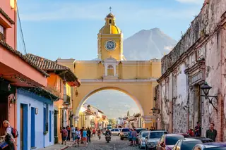 Yellow arch and clock tower above a cobbled street lined with colorful buildings, parked cars and small groups of pedestrians, with a volcano rising behind.