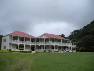 A white two-storey house with wraparound verandas and red roofs, behind a lawn and backed by trees.
