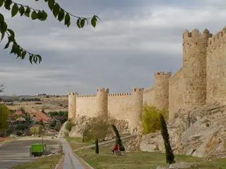 Crenellated stone towers follow a rocky slope above a paved path and a few slender trees.