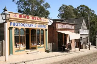 Colorful historic-style shopfronts with verandas, a bakery sign and a timber auction room along an unpaved street.
