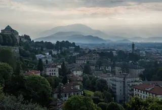 Layers of wooded hills rise behind red-roofed houses and green gardens under a hazy morning sky.