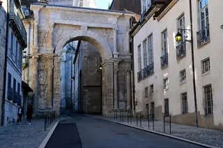 A tall pale-stone arch with carved reliefs spans a quiet narrow street between historic houses in Besançon.