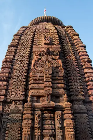 Intricate bands of figures, floral latticework and projecting carved stone details rise up a temple tower beneath blue sky.