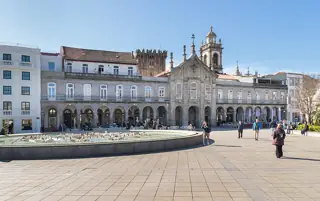 A long stone arcade and central church facade overlook a broad paved square and a low fountain basin.