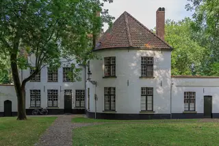 A rounded white facade and tiled roofs stand behind a lawn, footpath and leafy trees.
