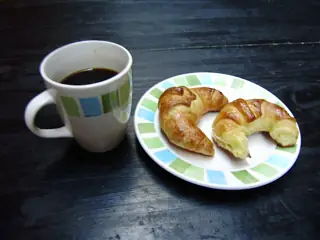 A mug of black coffee and two glossy crescent-shaped medialunas on a small plate on a dark wooden table.