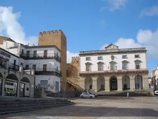 A pale town hall with an arcaded ground floor stands beside stone towers across an open paved square.