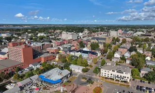 Colorful low-rise buildings and a twin-spired stone church among tree-lined streets beneath a blue, cloud-filled sky.