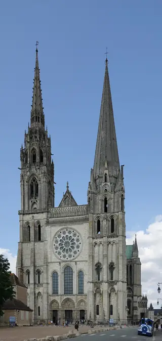 Two differently shaped stone spires rise above a rose window and three portals beneath blue sky.