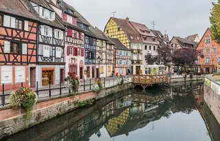 Colorful timber-framed houses and flower-lined banks reflected in a quiet canal beneath an overcast sky.