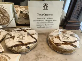 Two round powdered cakes in clear wrapping beside a Torta Cremona sign in a pastry-shop display.