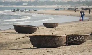 Round woven basket boats resting on sand beside the surf, with distant shore workers behind.