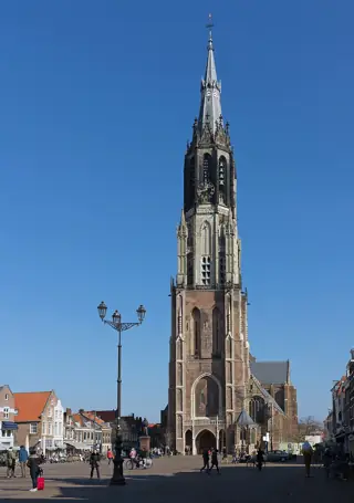 A tall stone clock tower and slender spire rise above people crossing a broad market square.