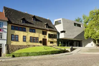 Yellow historic house beside an angular pale modern extension, with a lawn and cobbled square in front.