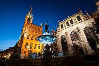 A brightly lit stone fountain and bronze Neptune stand between a tall orange-lit town hall and the pale arched façade of Artus Court beneath a deep blue sky.