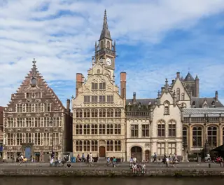 Ornate stepped-gable facades and a clock tower stand above a stone quay where small groups sit beside the water.