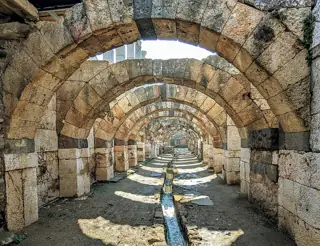 Rows of broad stone arches recede over a narrow water channel and a sunlit gravel floor.