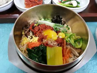 A metal bowl of rice, vegetables and egg with side dishes on a blue table covering.