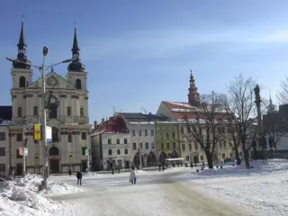 Snow across a broad square with a twin-towered church, colorful buildings, bare trees and distant pedestrians.