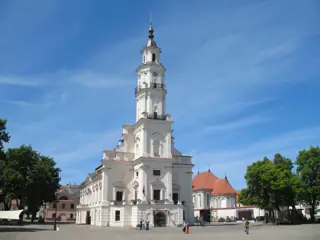 A tall white clock tower rises above a white town hall facing an open paved square with small groups of visitors.