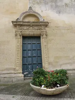 Ornate stone portal surrounding a weathered blue door, with a large planted bowl in the foreground.