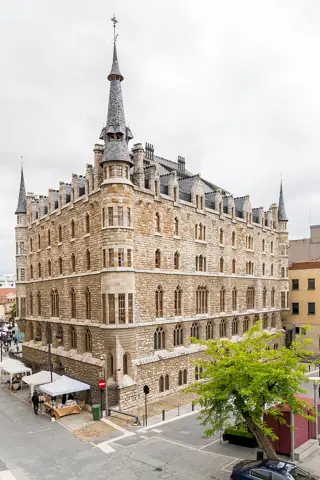 A turreted pale-stone building with steep slate roofs overlooks a paved square with small market tents.