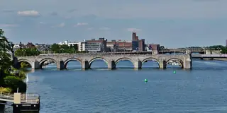 Stone bridge arches cross blue river water, with small walkers on the deck and city buildings beyond.