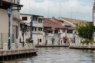 Low riverside houses with red and grey roofs line a curved walkway beside brown water and blue lamps.