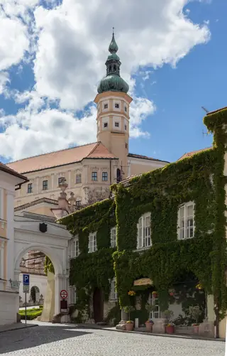 A copper-domed tower rises behind pale castle walls, an entrance arch and an ivy-covered building beside a cobbled street.