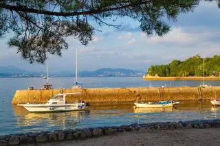 Small boats float near a stone breakwater and pine-lined shoreline, with distant hills and the mainland across the water.