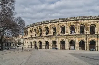 Weathered stone arena arcades under gray clouds, with a few visitors across the square.