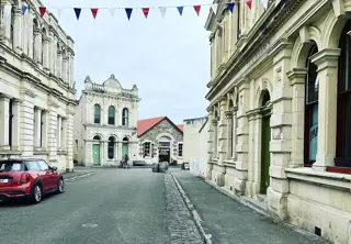 Historic limestone facades line a quiet street with bunting, a red car and cyclists near the far end.