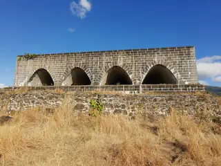 Four pointed openings in a basalt-stone fort wall above dry grass, against blue sky.