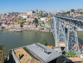 Iron arch of Dom Luís I Bridge above tiled and metal roofs, with Porto rising across the river.