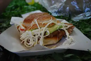A sesame-topped cemita filled with shredded white cheese, avocado and breaded meat, resting on a paper-lined tray.
