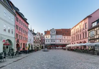 Quiet cobbled square between pastel and half-timbered facades, with café tables and parasols.