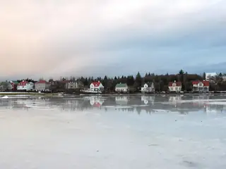 Broad frozen pond reflecting a row of houses beneath a pale blue-gray winter sky.
