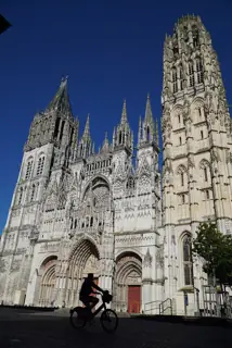 Ornate stone cathedral facade and two towers above a sunlit square, with a cyclist in foreground shadow.