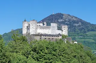 White fortress walls rise above green trees, with scaffolding around one section and a wooded mountain behind.