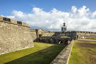 Sunlit stone fortifications and a square lighthouse rise above a grassy moat beneath a blue, clouded sky.