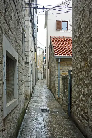 Wet stone paving runs between narrow residential facades beneath overhead wires.