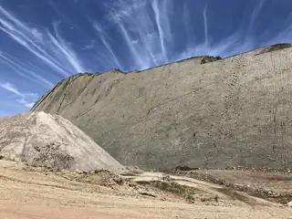 Tall pale rock face crossed by dinosaur tracks above a dusty quarry floor beneath wispy clouds.