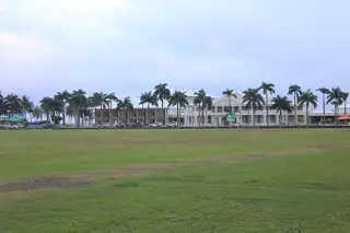 A broad grass field faces a white hotel and palms beneath pale gray clouds.