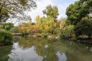 Orange and green trees surround a pond with a low stone bridge and reflections of garden buildings.