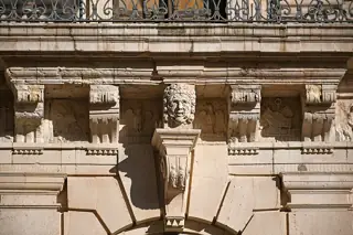 A carved stone mask, corbels and relief panels beneath an iron balcony railing.