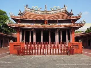 A red temple hall with tiled roofs, sculpted dragons and a red fence faces a stone-paved courtyard.