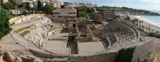 A wide view of a stone amphitheatre, church foundations and surrounding park beside the coast.
