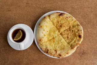 Round cheese bread on a plain white plate beside lemon tea in a white cup on a brown table.